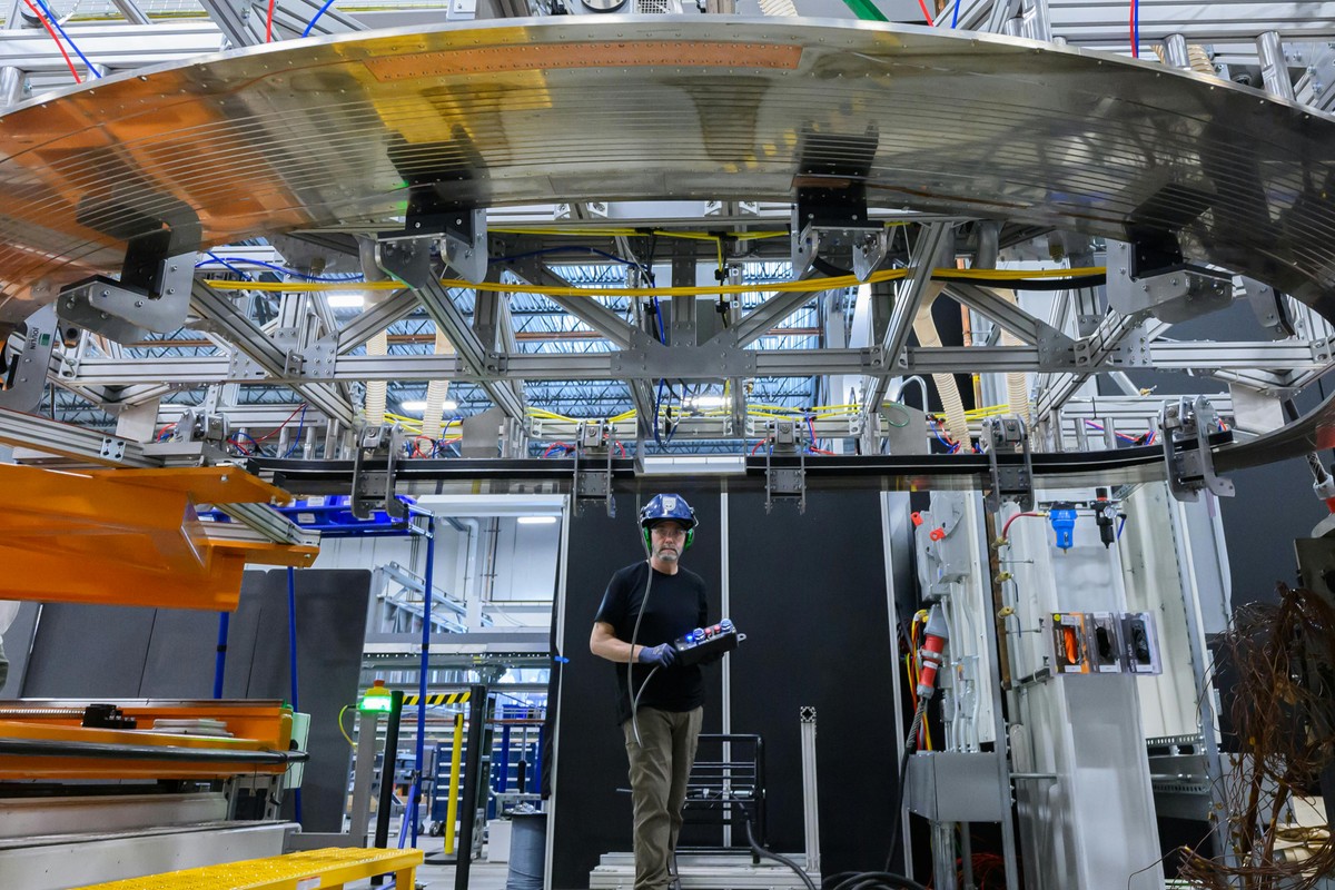 A man, wearing a hardhat and holding a crane control box, stands behind a D-shaped steel plate with embedded copper-colored electrical channels that's been hoisted above a factory floor.