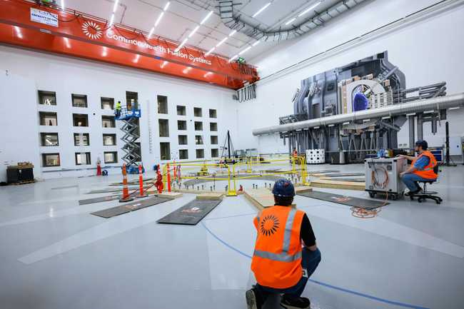 Two people in orange safety vests work on the floor of a spacious hall with a crane above and an illustration of a tokamak to generate fusion power on one wall. Another wall, pierced by rectangular holes for diagnostic equipment. A third person stands on a mobile scissor lift with its platform extend upward. 