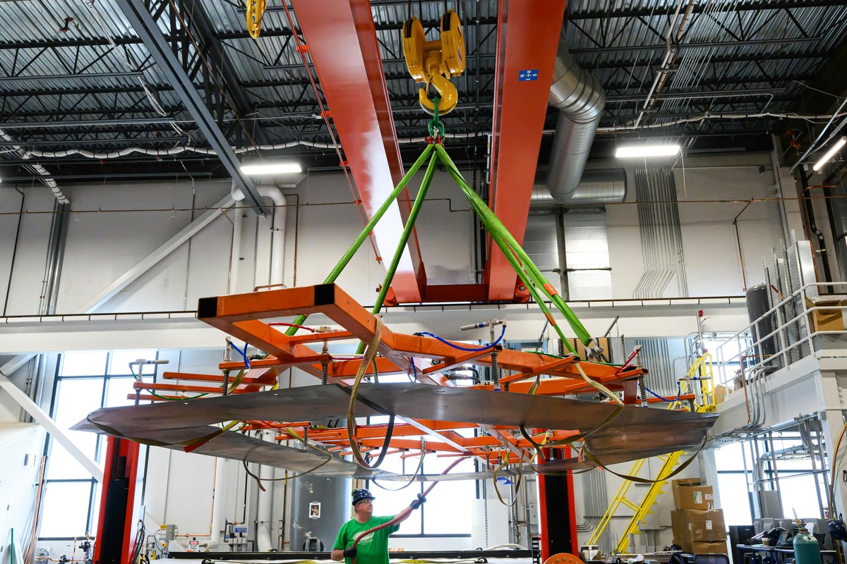 A person in a hard hat stands near a D-shaped copper plate attached to an orange metal frame that's suspended from a factory crane