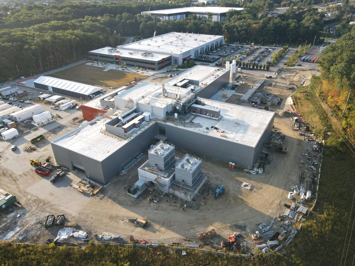 An aerial view of a white-roofed, plus-shaped building with construction equipment and material nearby, another large building behind it, and a green forest surrounding it