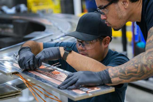 Two people wearing safety glasses feeding copper-colored tape into grooves on a steel plate in the factory