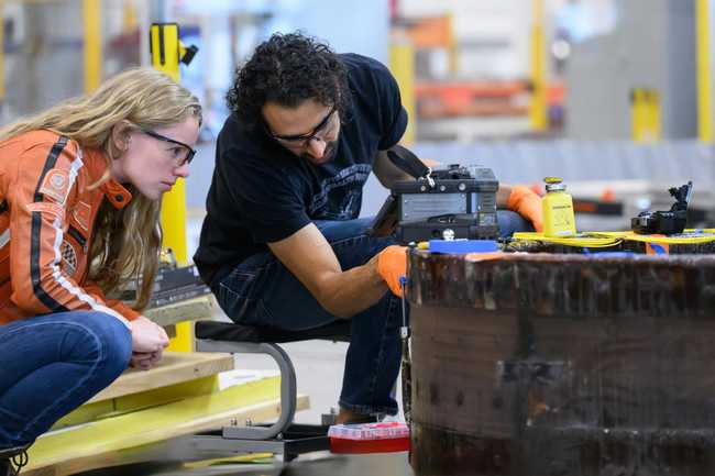 Two people wearing safety glasses use electronic instruments to probe a cylindrical metallic structure on a factory floor.