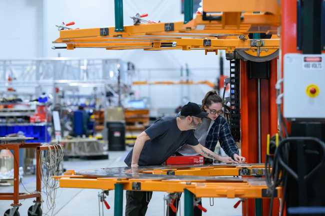 Two wokrers wearing safety glasses adjust a metal plate resting on an orange stand on a factory floor