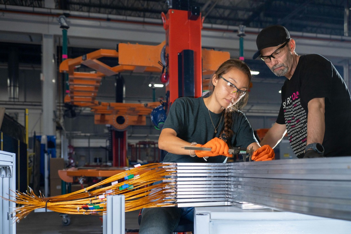 Two people measuring a stack of precisely aligned steel plates, each with a bundle of copper-colored cable connectors, in a factory setting