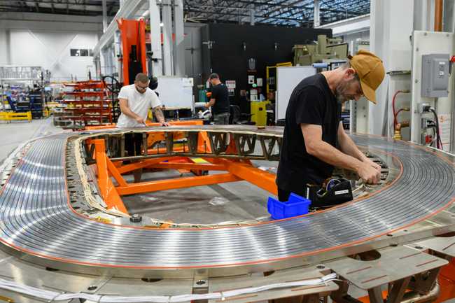 Two people work on wiring connections on a D-shaped steel plate that's grooved with parallel, concentric channels.